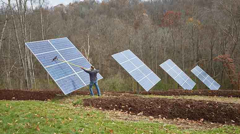 Man outside cleaning solar panels at his home in a field with woods in background